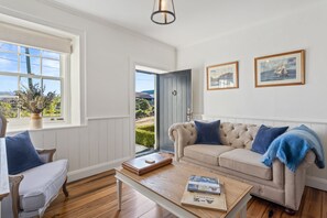 A living room with a beige sofa, a wooden coffee table, and a window with a view of the outdoors. - Lythgo's Row Colonial Cottages (Pontville)