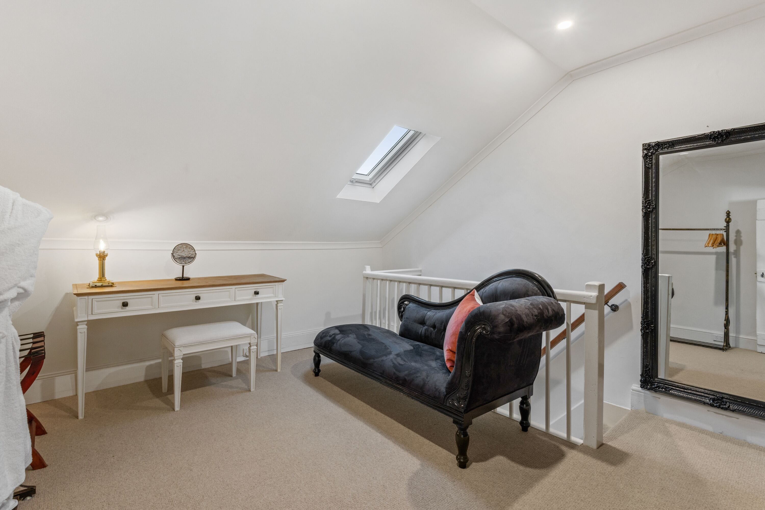 A bedroom with a sloped ceiling, a desk, a bench, a dark sofa, a large mirror, and a small window.