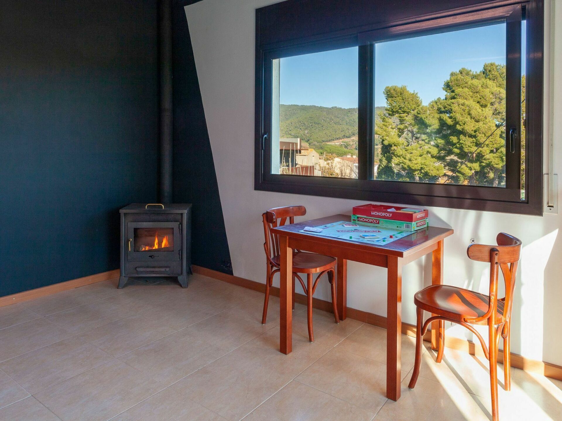 A cozy room with a wood-burning stove, a table with Monopoly board games, and a window overlooking a green landscape.