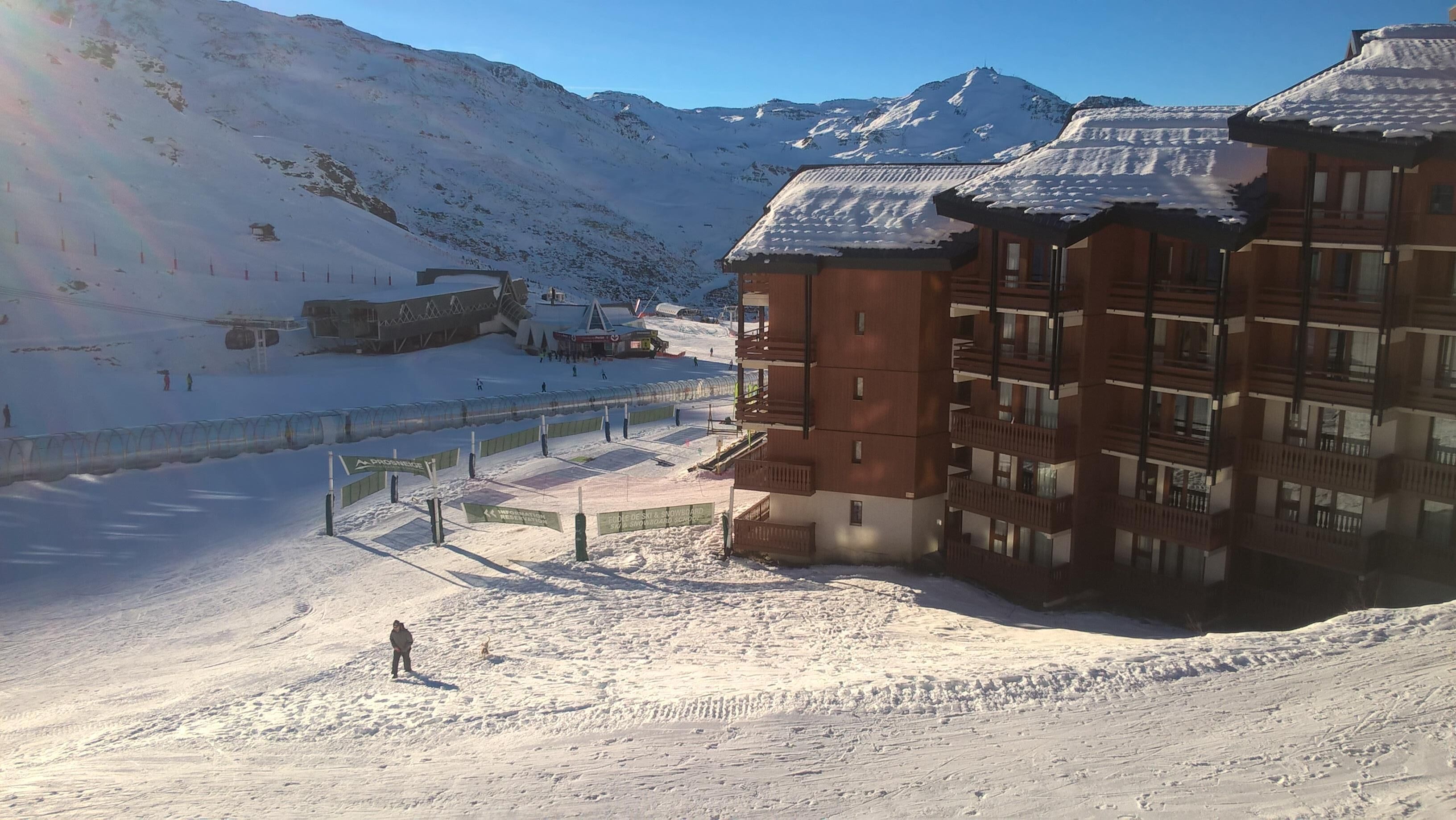 A snow-covered ski resort with a wooden building featuring balconies, a ski lift, and a clear view of the mountains.