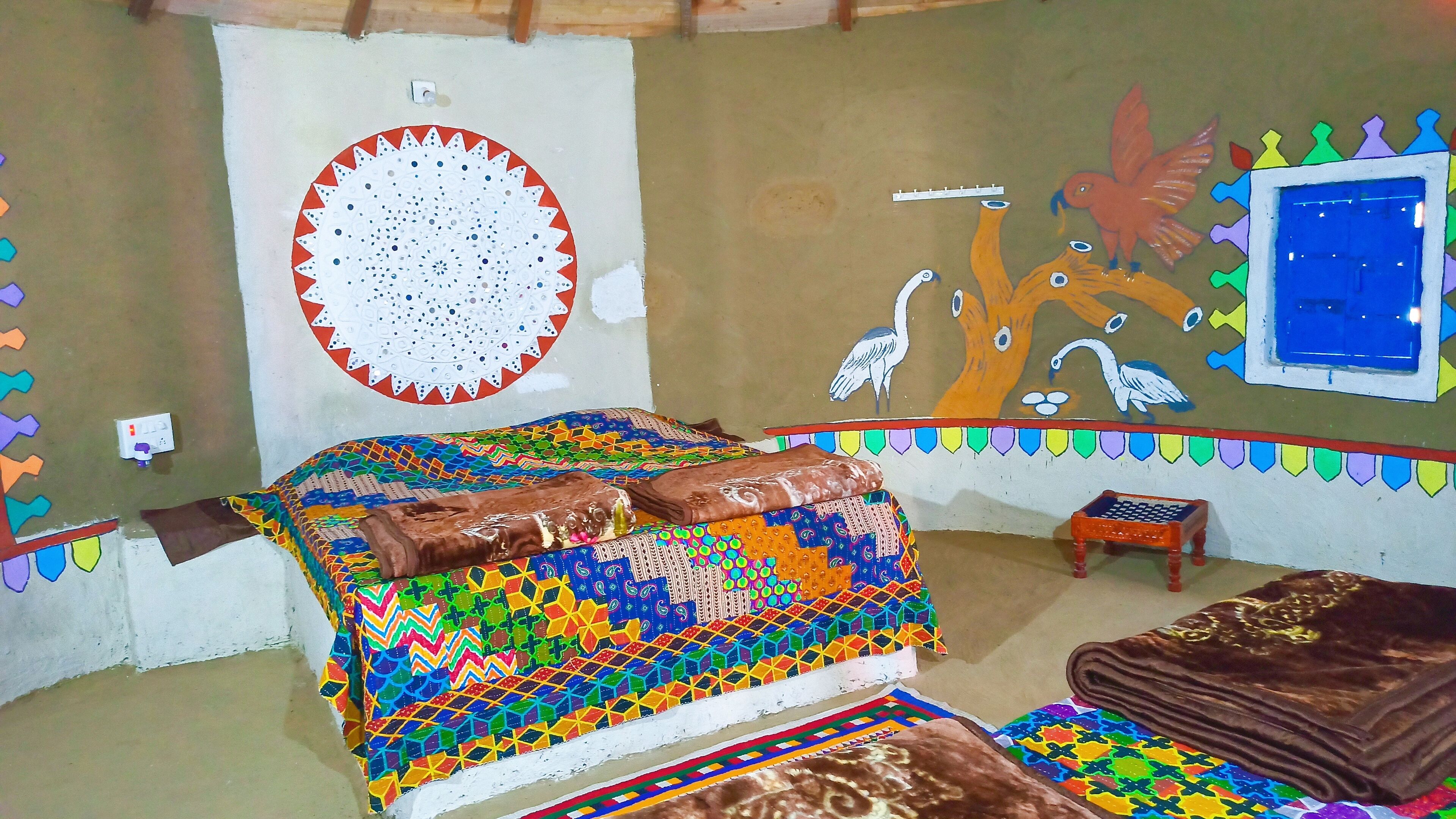 Una habitación con una colcha de colores, una pared con un diseño circular y un mural de pájaros y un árbol.