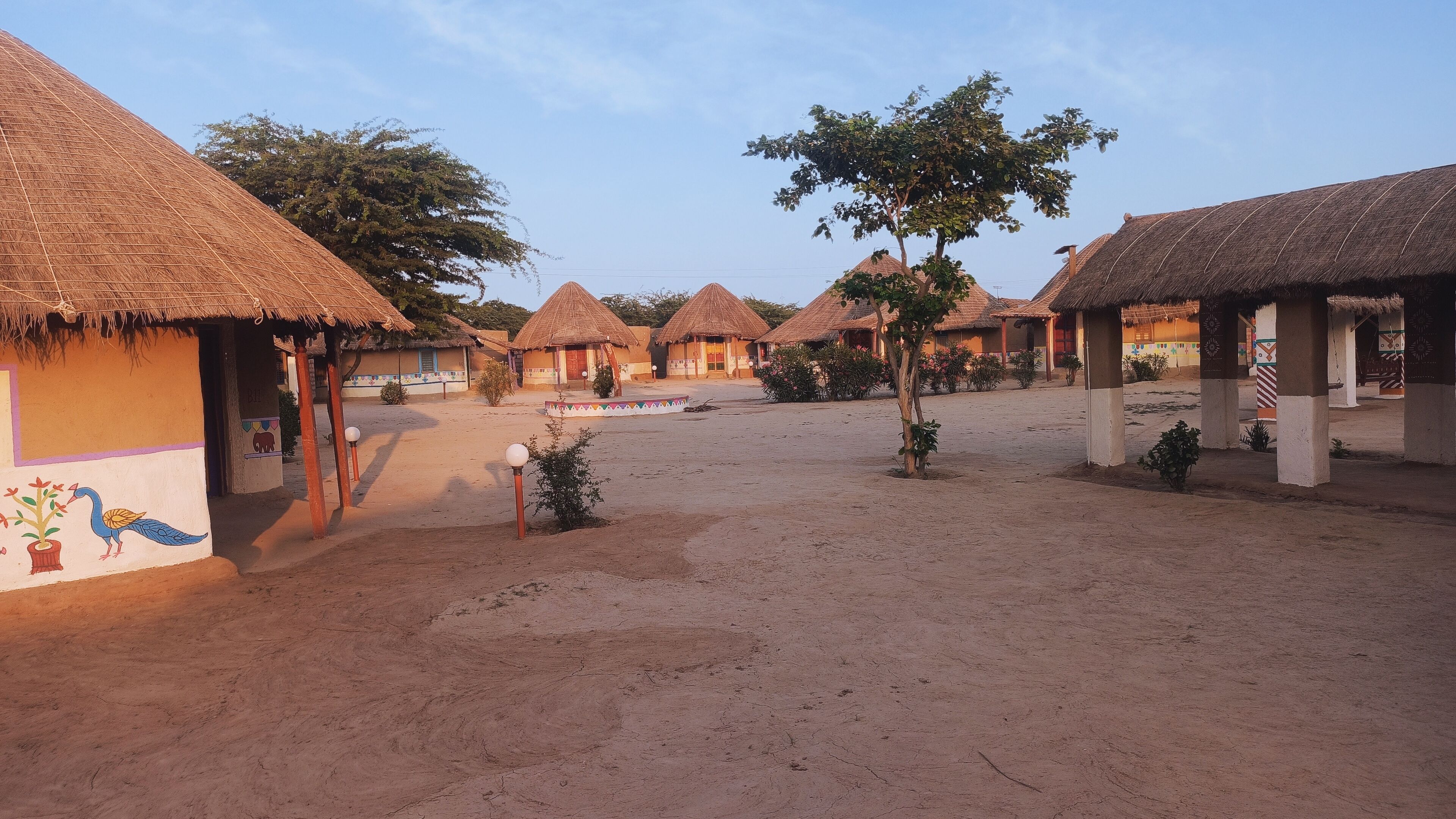A village with thatched-roof huts, a sandy ground, and a few trees.