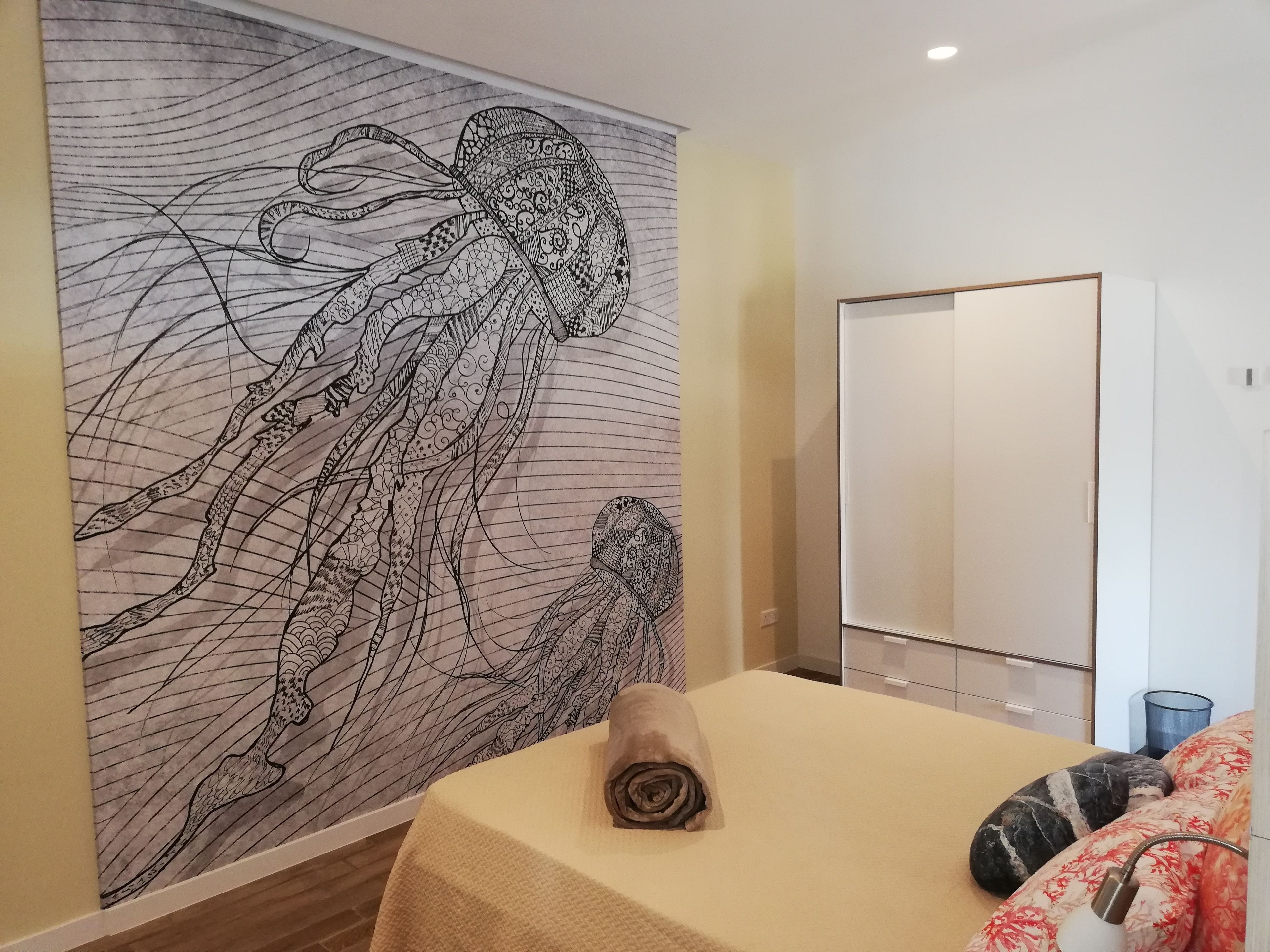 Una habitación con un tapiz de pared grande, una cama con una toalla enrollada, un armario y una mesita de noche con lámpara.