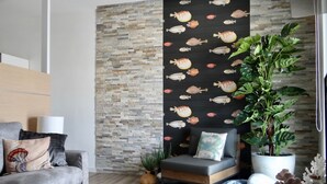 Una sala de estar moderna con un sofá gris, una pared negra con motivos de peces, una planta verde alta y una pared con textura de piedra.