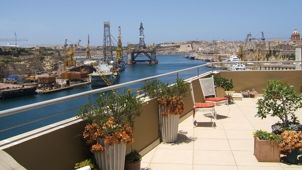 Una terraza con plantas en macetas y mobiliario exterior, con vista a un puerto con barcos y grúas.