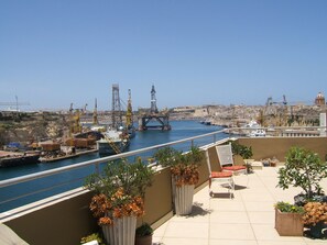 A terrace with potted plants and outdoor seating overlooking a harbor with ships and cranes. - Three Cities Apartments (Cospicua)