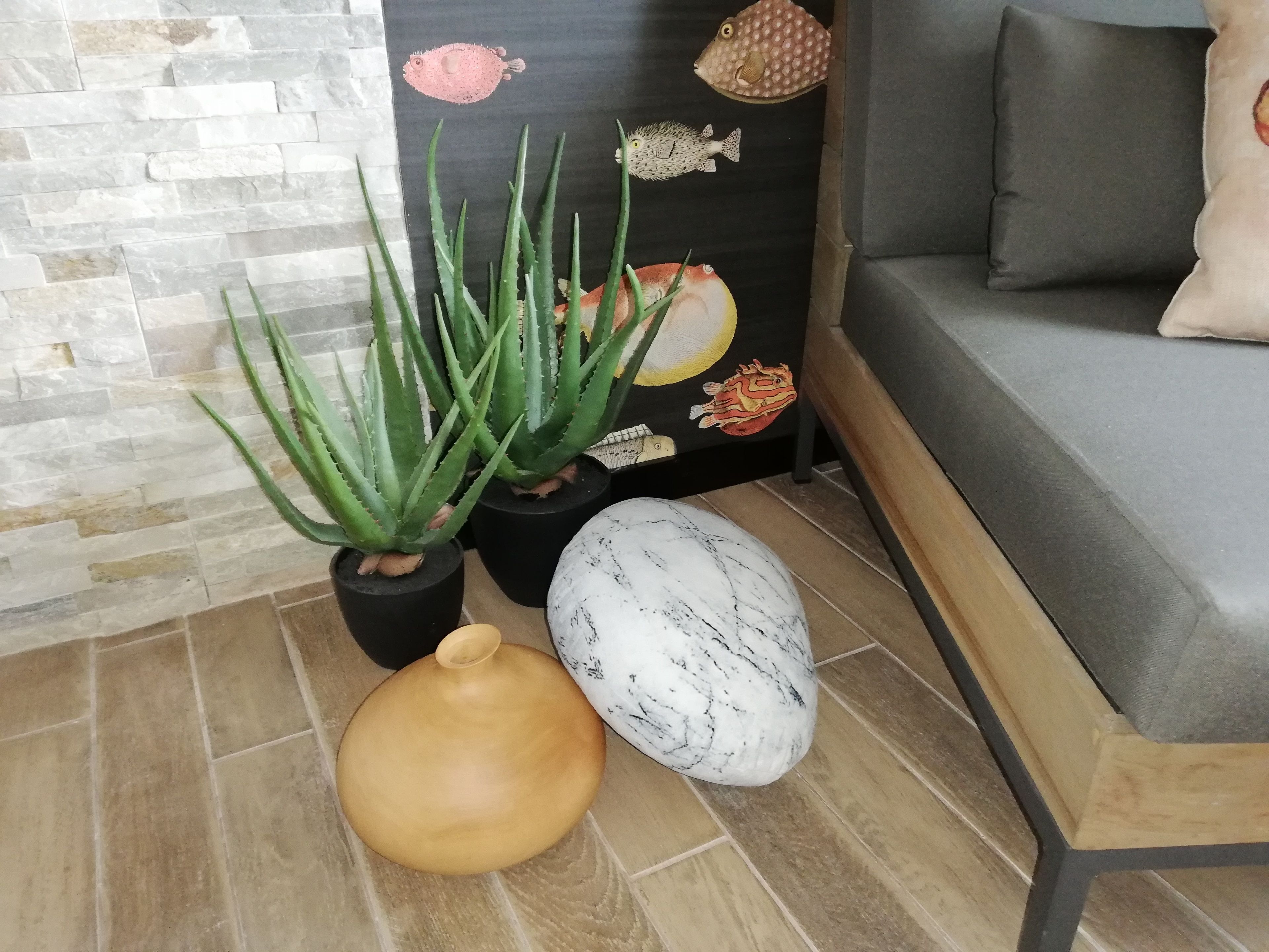 Una esquina de una habitación con una planta de aloe vera en maceta, un cuenco de madera y una escultura de mármol en el suelo.