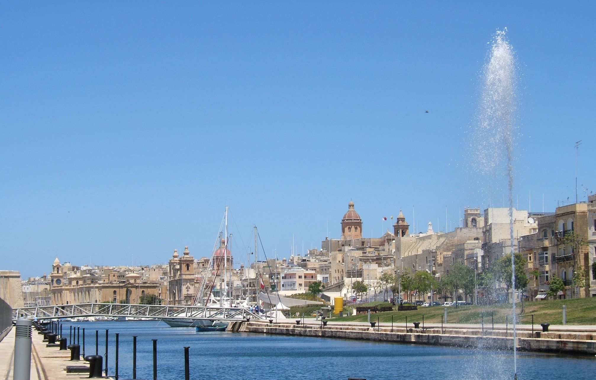 Une vue urbaine sur le front de mer, avec une fontaine imposante, des bâtiments historiques et un port de plaisance rempli de bateaux.