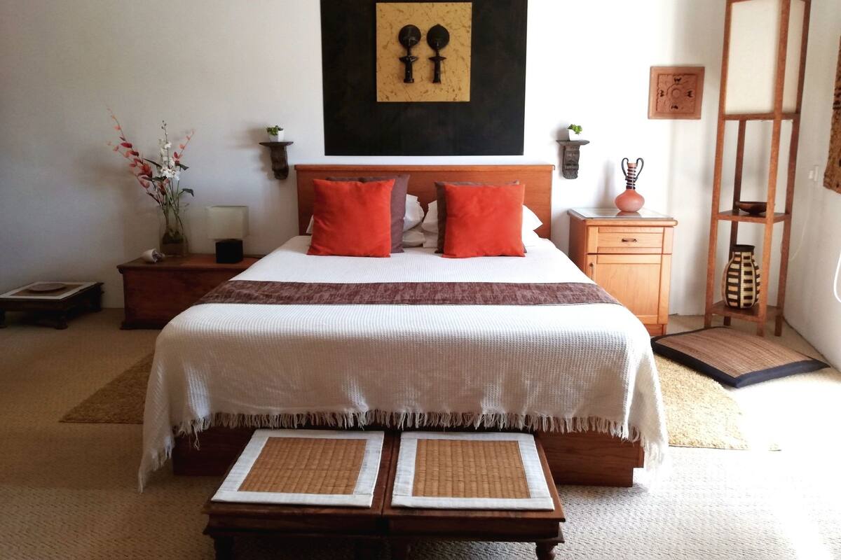 A neatly arranged bedroom with a large bed, two bedside tables, a wooden nightstand, a small shelf, and a decorative wall piece.