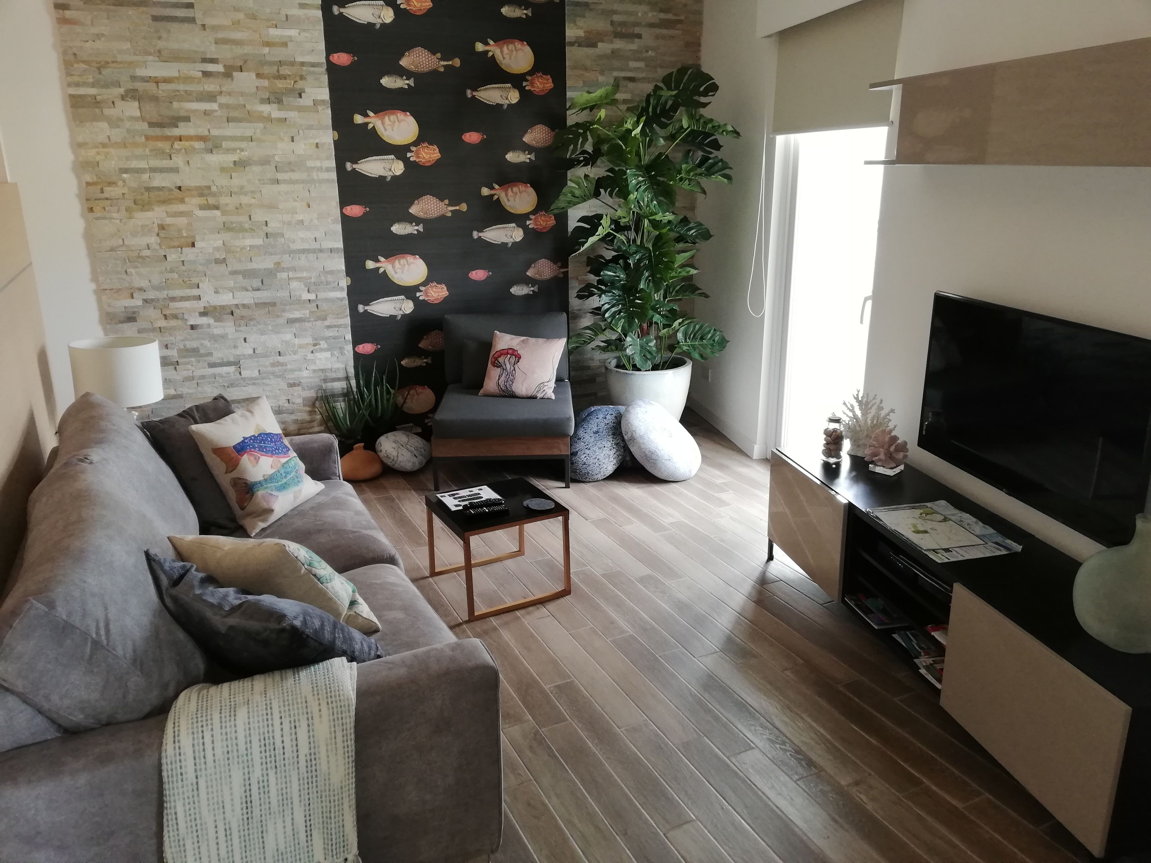 A modern living room with a grey sofa, a black accent wall with fish patterns, a large potted plant, a flat-screen TV, and a wooden coffee table.