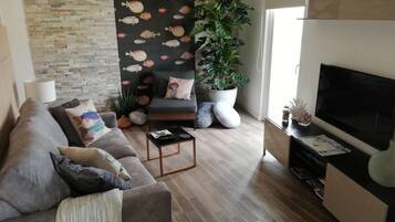 A modern living room with a grey sofa, a black accent wall with fish patterns, a large potted plant, a flat-screen TV, and a wooden coffee table.