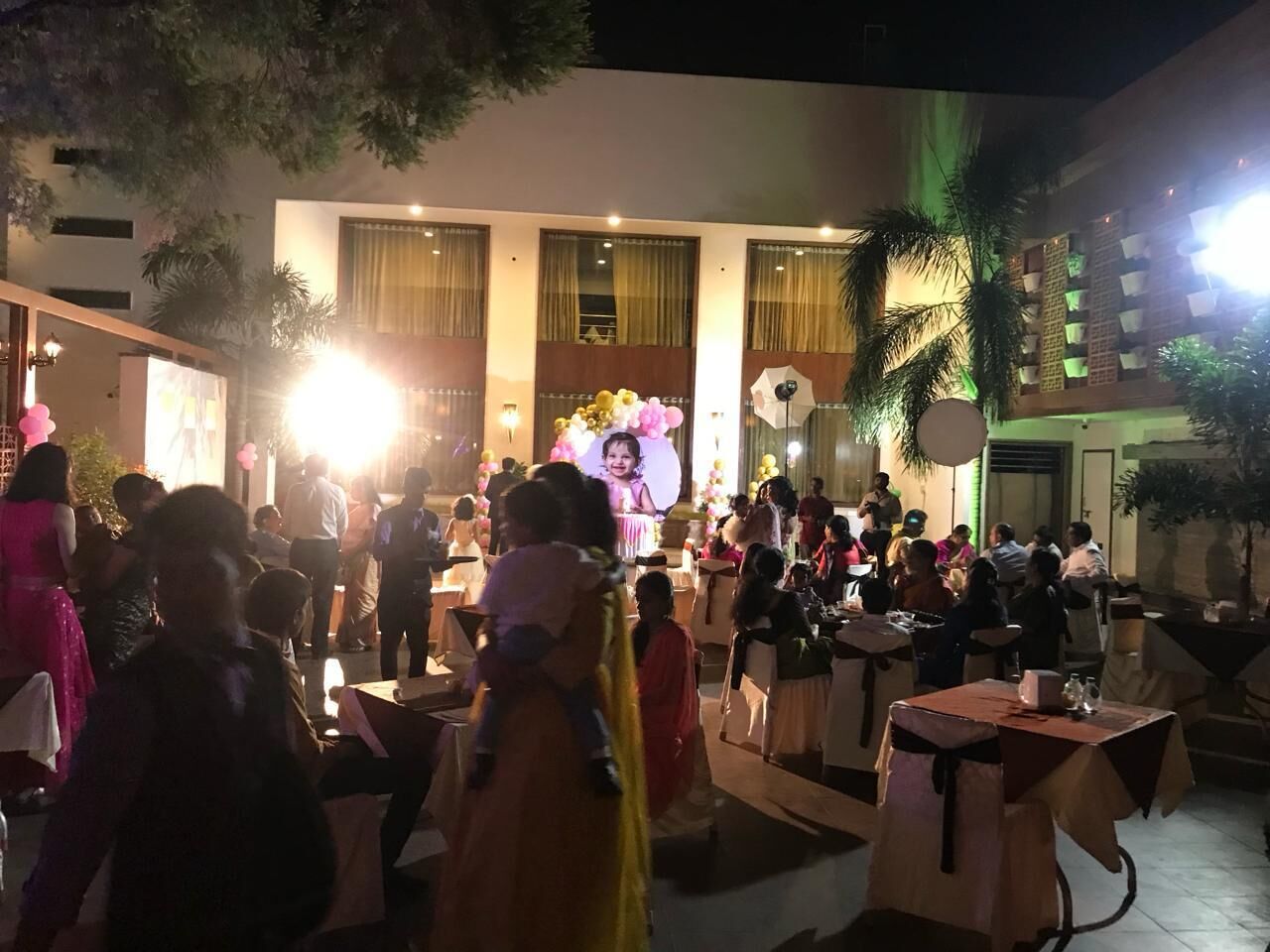 Evento al aire libre con mesas y sillas dispuestas para los asistentes, un escenario con fondo y una multitud de personas.