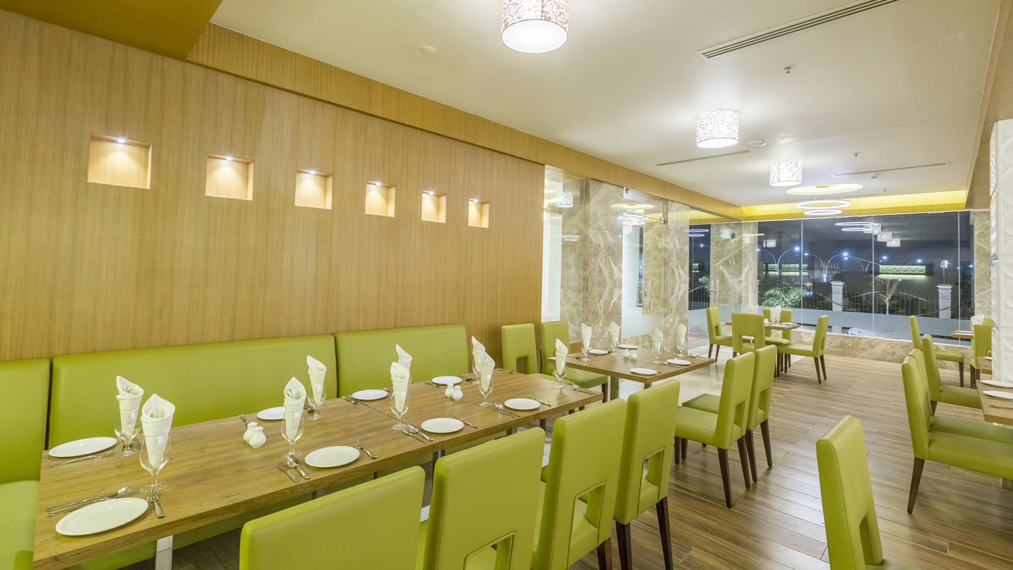 Un restaurante con largas mesas de madera, asientos tapizados verdes y luminarias modernas.