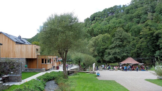 A riverside area with a wooden building, a small bridge, and a gathering of people near a pavilion. - Cozy chalet in a quiet, green area (Rendeux)