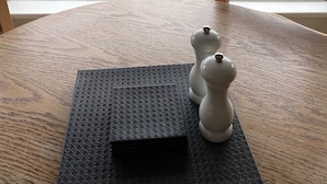 Een kamer met een groot raam met uitzicht op een woonwijk, een houten stoel en een peper- en zoutstelset op een gestructureerde mat.