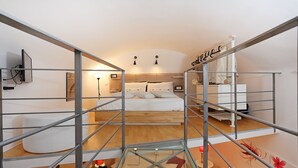 Una habitación de hotel moderna con techo de cristal, suelo de madera, cama con ropa blanca y una pequeña zona de estar con un cojín rojo.