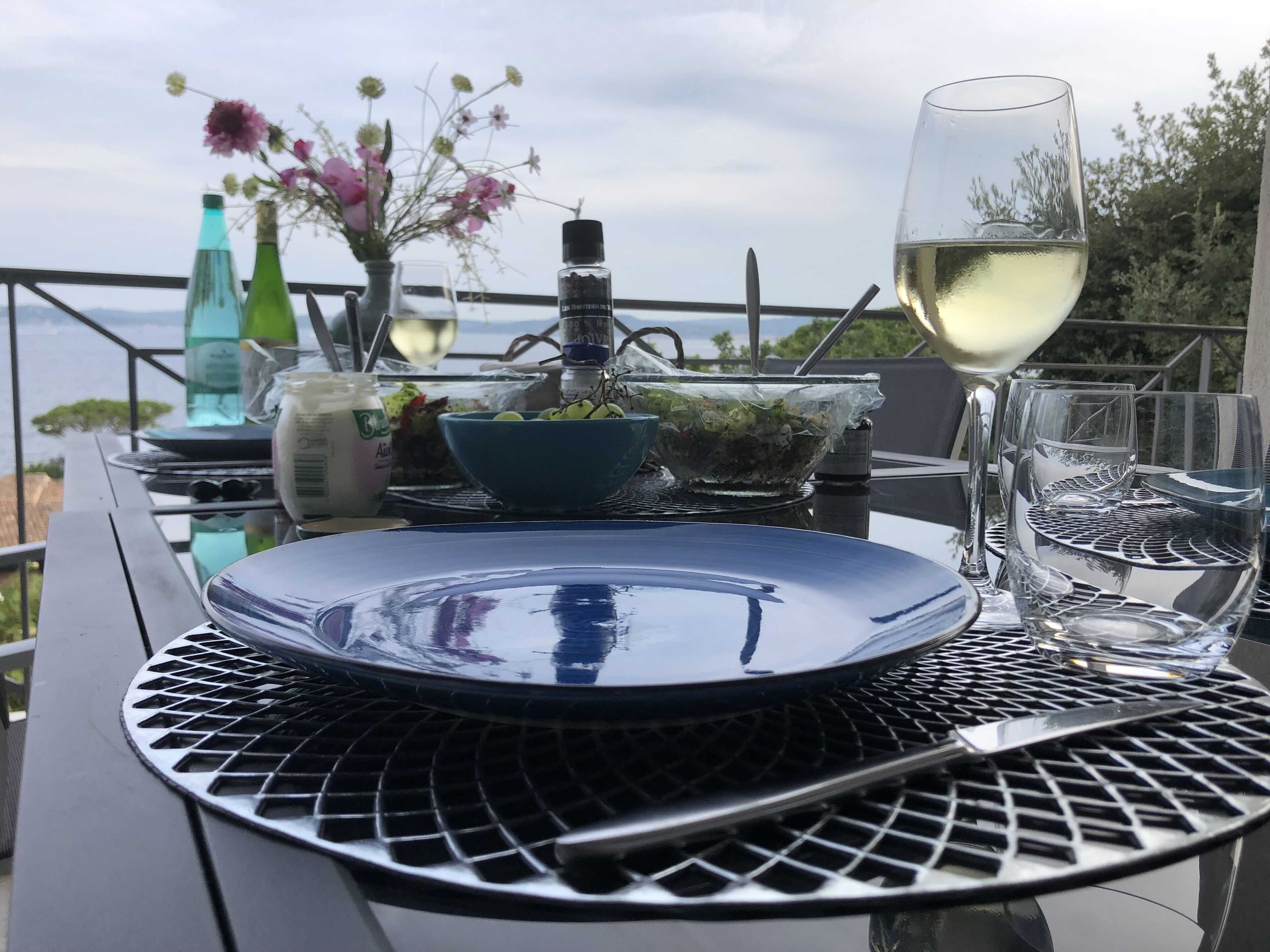 A table set with a blue plate, a glass of white wine, a bowl of salad, and a bottle of dressing.