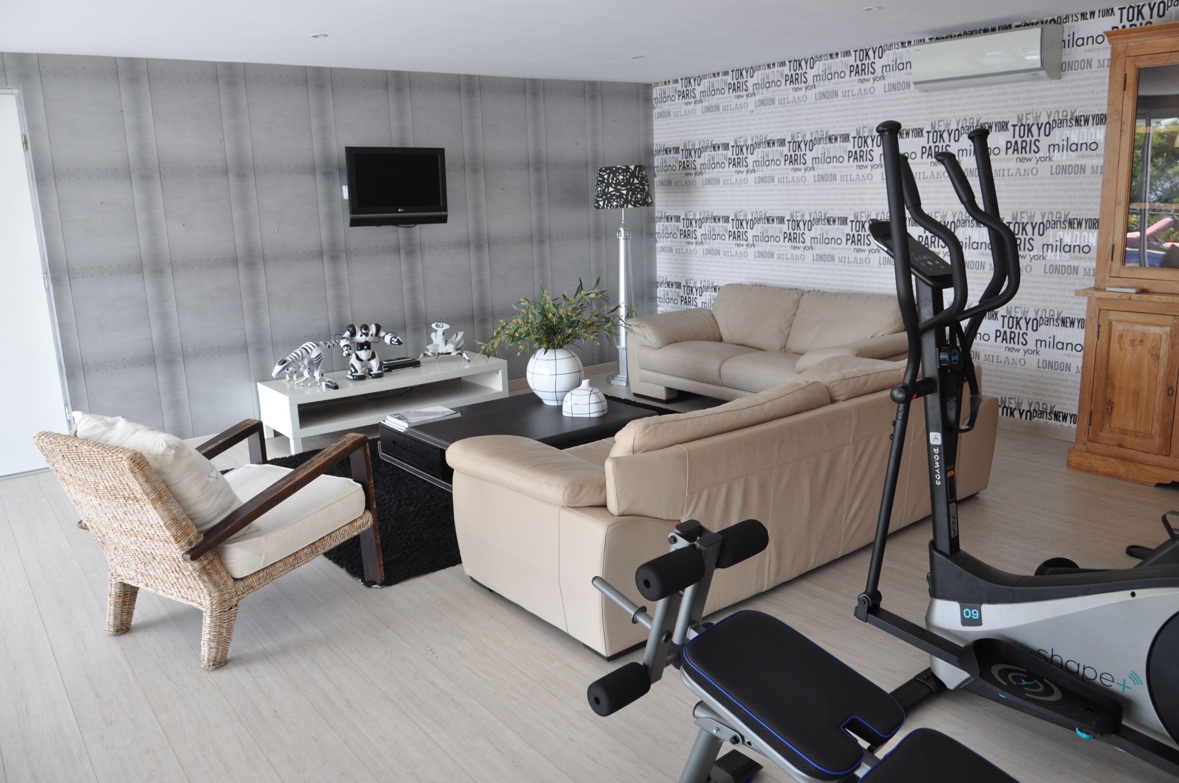 A modern living room with a sofa, a chair, a TV, a small table with decorative items, and a stationary exercise bike.