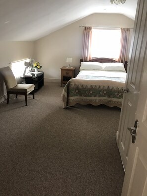 Une chambre à coucher avec un lit, une chaise, une table de chevet, un ventilateur et une fenêtre avec des rideaux.