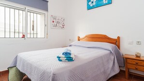 A bedroom with a bed, a window with bars, a wall with a floral painting, and a nightstand with a lamp. - Centro Vejer Apartment with Terraces and Beautiful Views (Vejer de la Frontera)