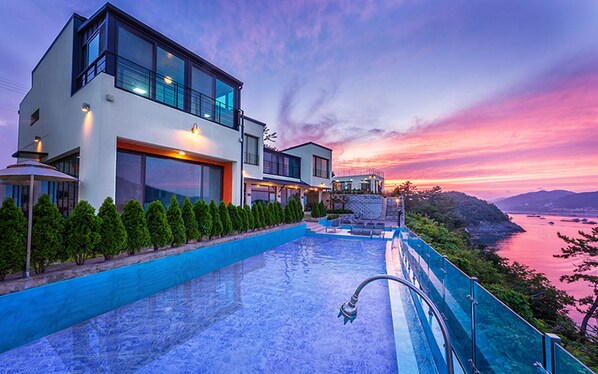 A modern two-story house with a swimming pool, large windows, and a view of the water and mountains at sunset. - Good Time Resort (Yeosu)