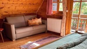 Ein gemütliches Cabin-Zimmer mit Holzbalkendecke, einer Couch, einem Bett mit karierter Bettwäsche und einem Fenster mit Vorhängen.
