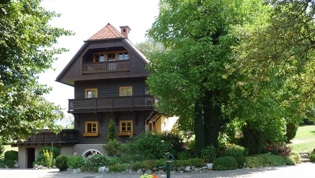 Ein zweistöckiges Holzhaus mit einem Balkon, umgeben von üppigem Grün und einem gepflasterten Einfahrt.