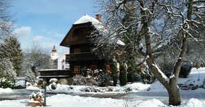 A traditional wooden house with a snow-covered roof, surrounded by snow-laden trees and a clear sky.