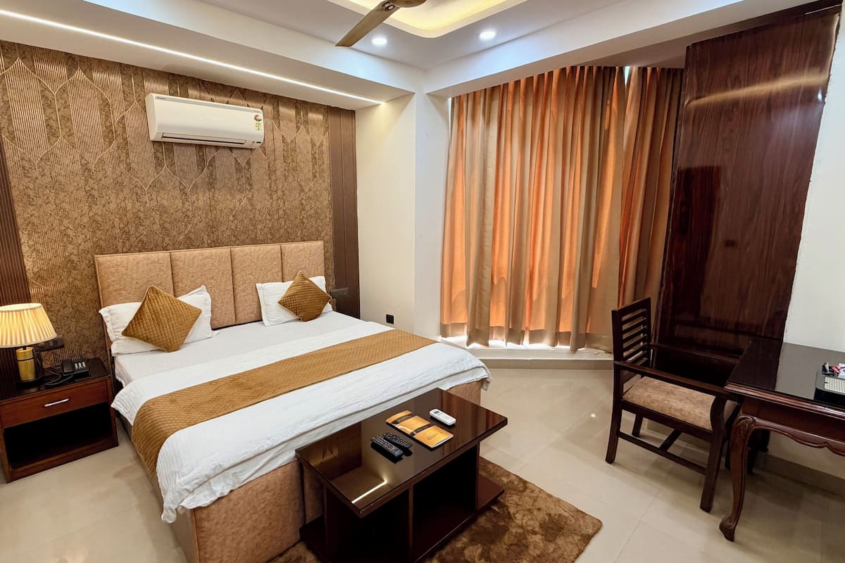 A hotel room with a bed, bedside table, lamp, chair, desk, and an air conditioning unit.