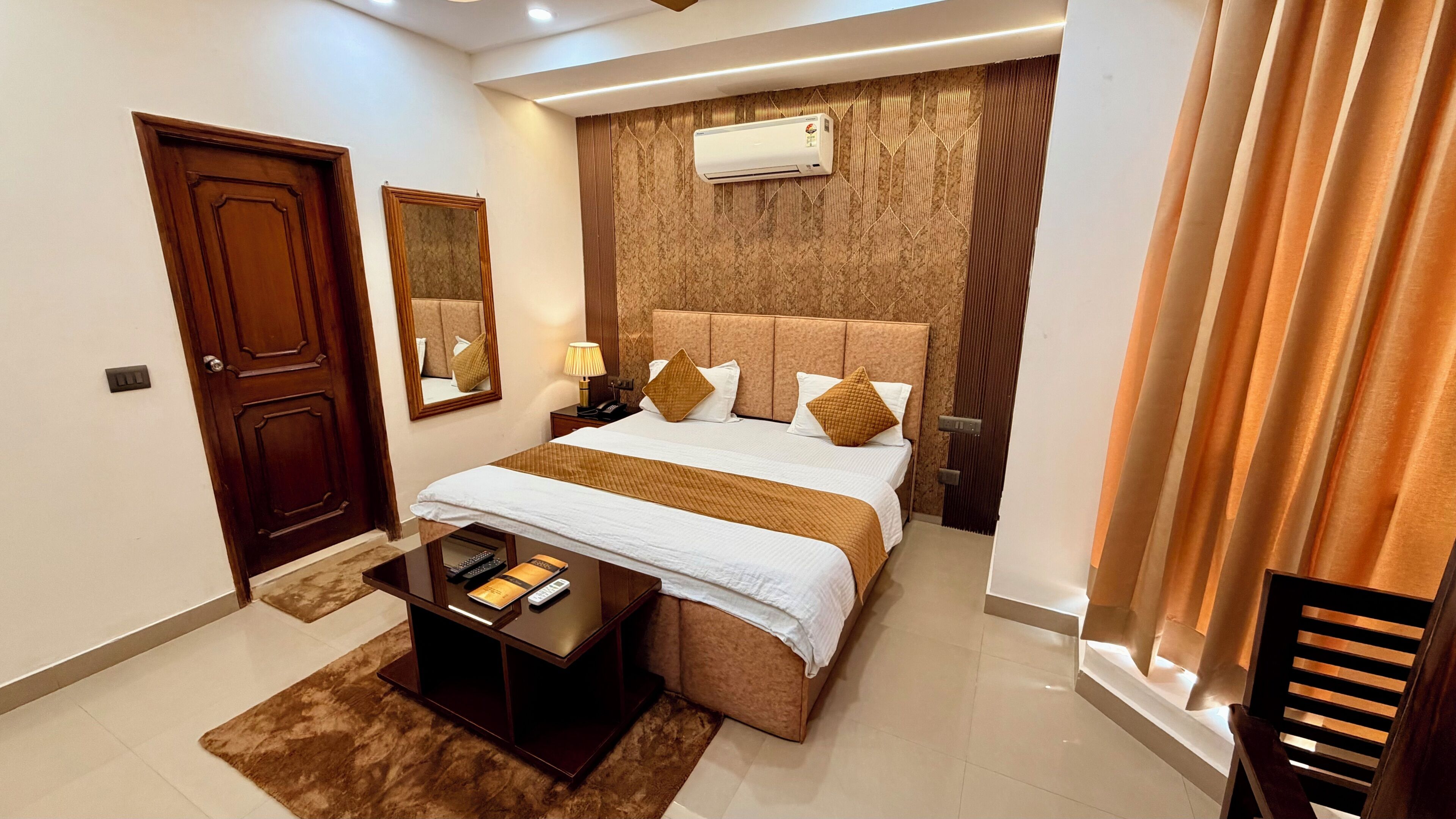 A hotel room with a bed, bedside table, lamp, air conditioning unit, and a door.