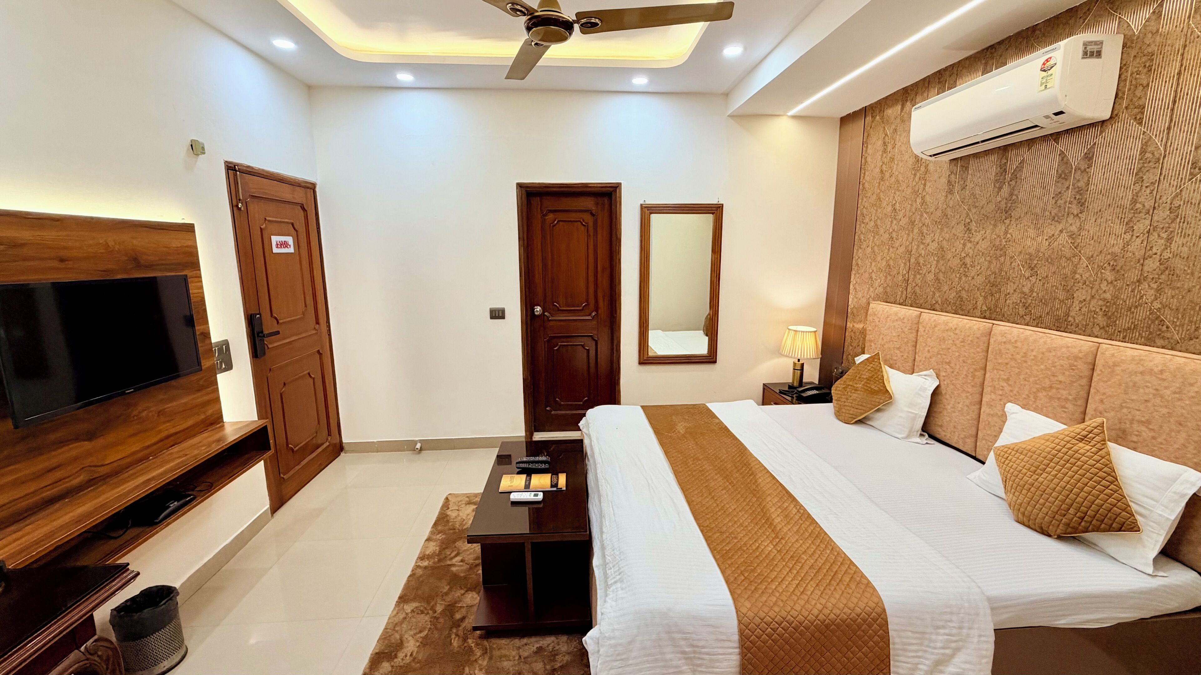 A hotel room with a bed, TV, ceiling fan, air conditioning unit, and a mirror.