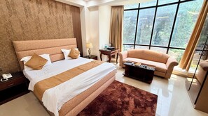 A bedroom with a large bed, a sofa, a coffee table, a nightstand, and a window with a view. - Kashish Residency - Premium Rooms (Noida)