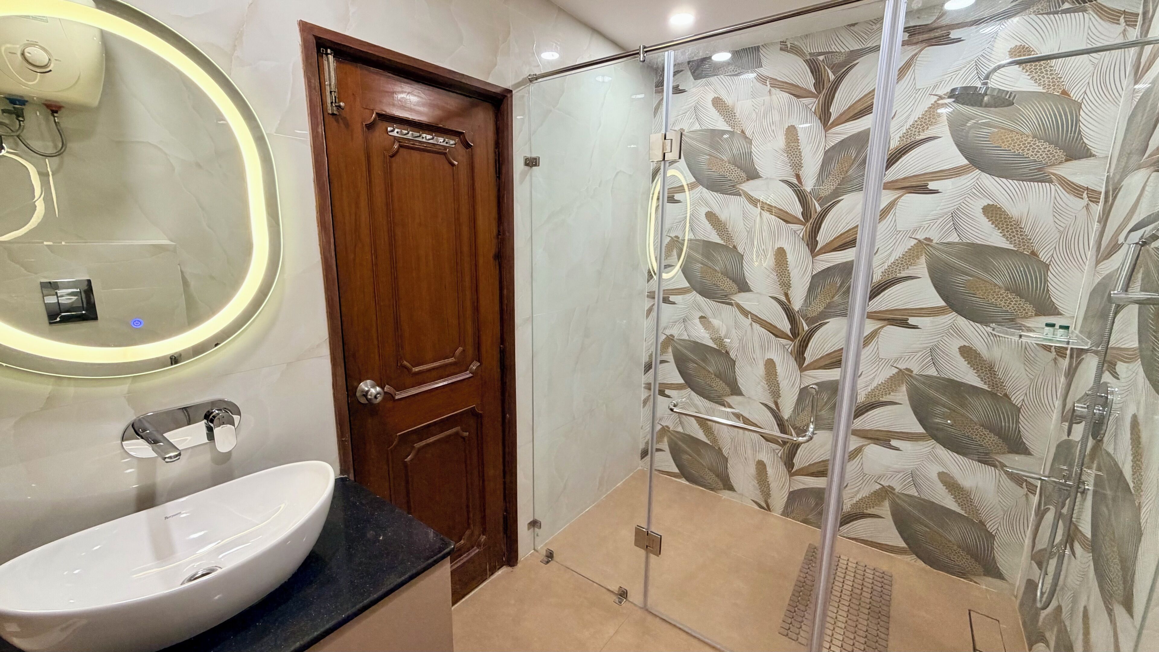 Un baño con puerta de madera, espejo redondo, lavamanos blanco y ducha con puerta de vidrio con diseño de hojas.