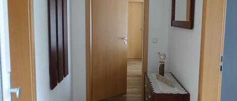 Un couloir étroit au plancher de bois, une commode en bois recouverte d’une housse en dentelle et un miroir sur le mur de droite.