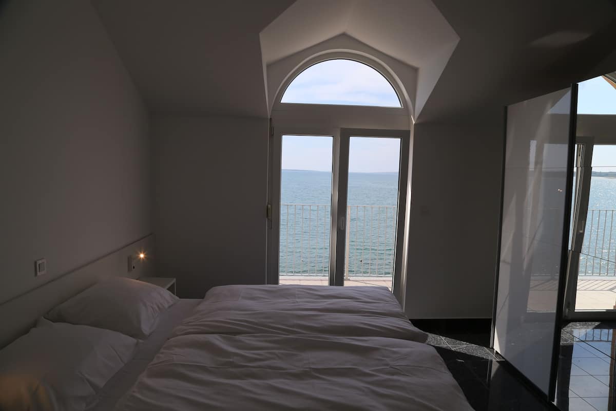 A bedroom with a large bed, a nightstand, and a view of the sea through a glass door.