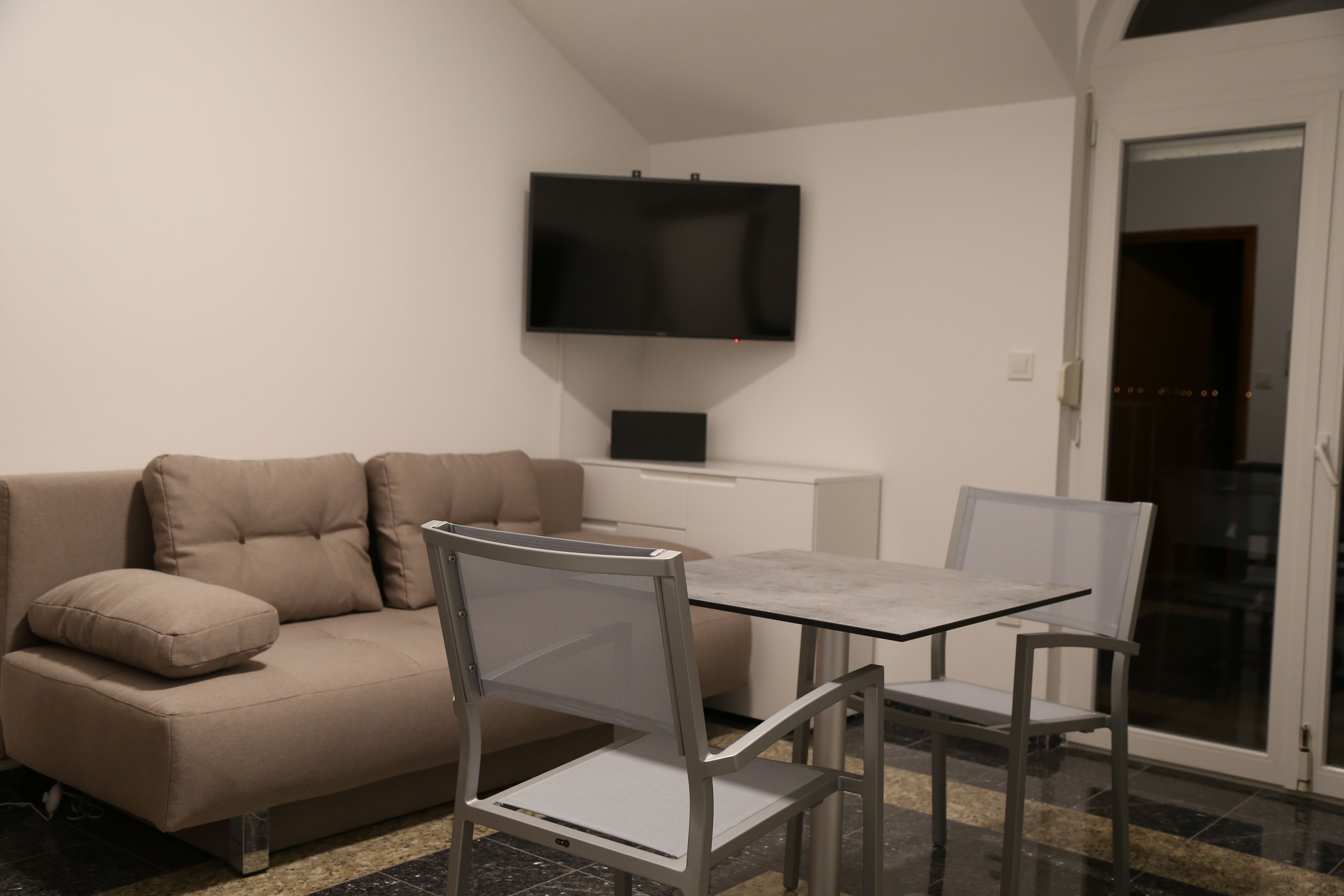A modern living room with a sofa, a glass-top dining table, and two chairs. A flat-screen TV is mounted on the wall above the sofa.