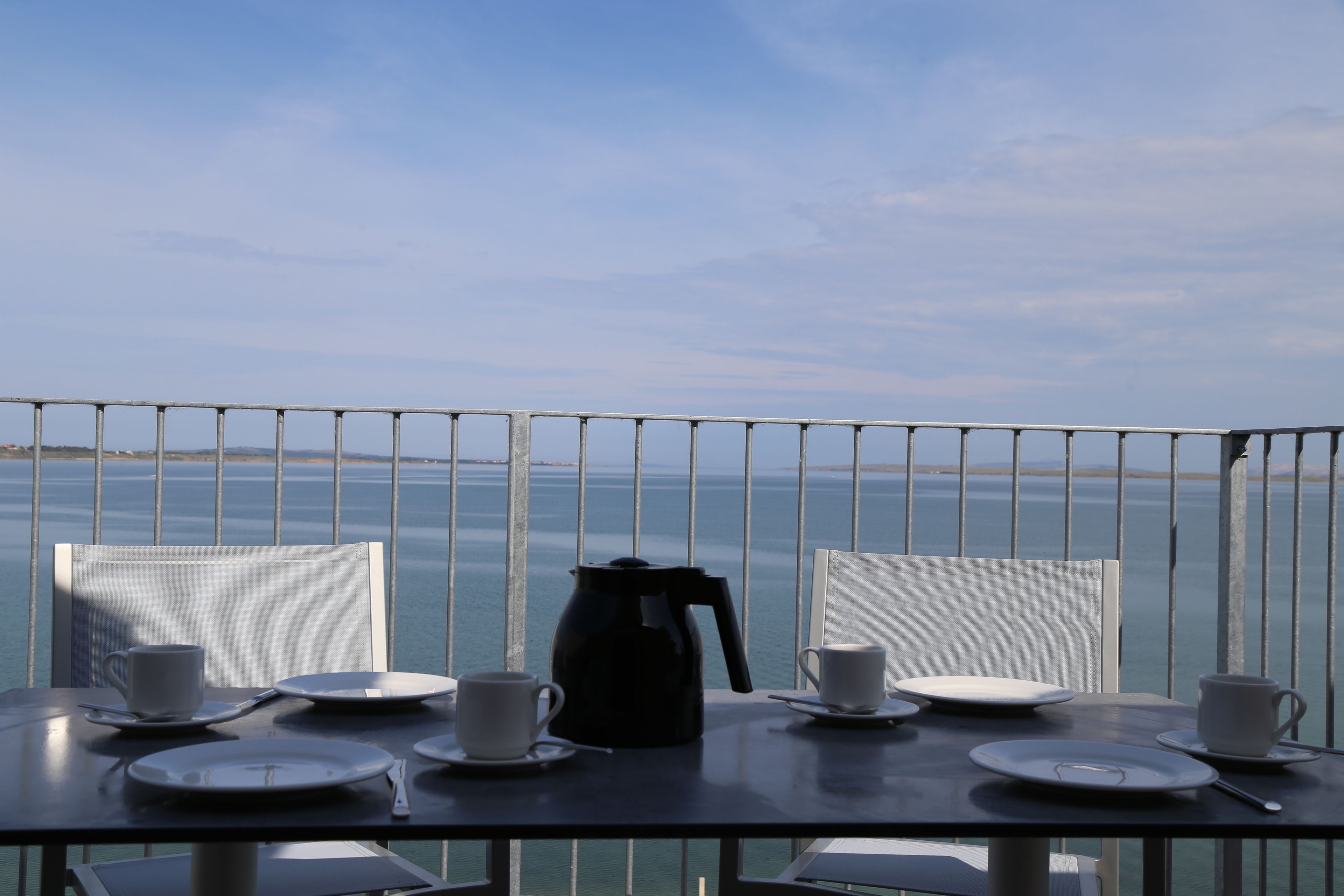 Una mesa con platos, tazas y una cafetera de café negro, frente a una vista al mar.