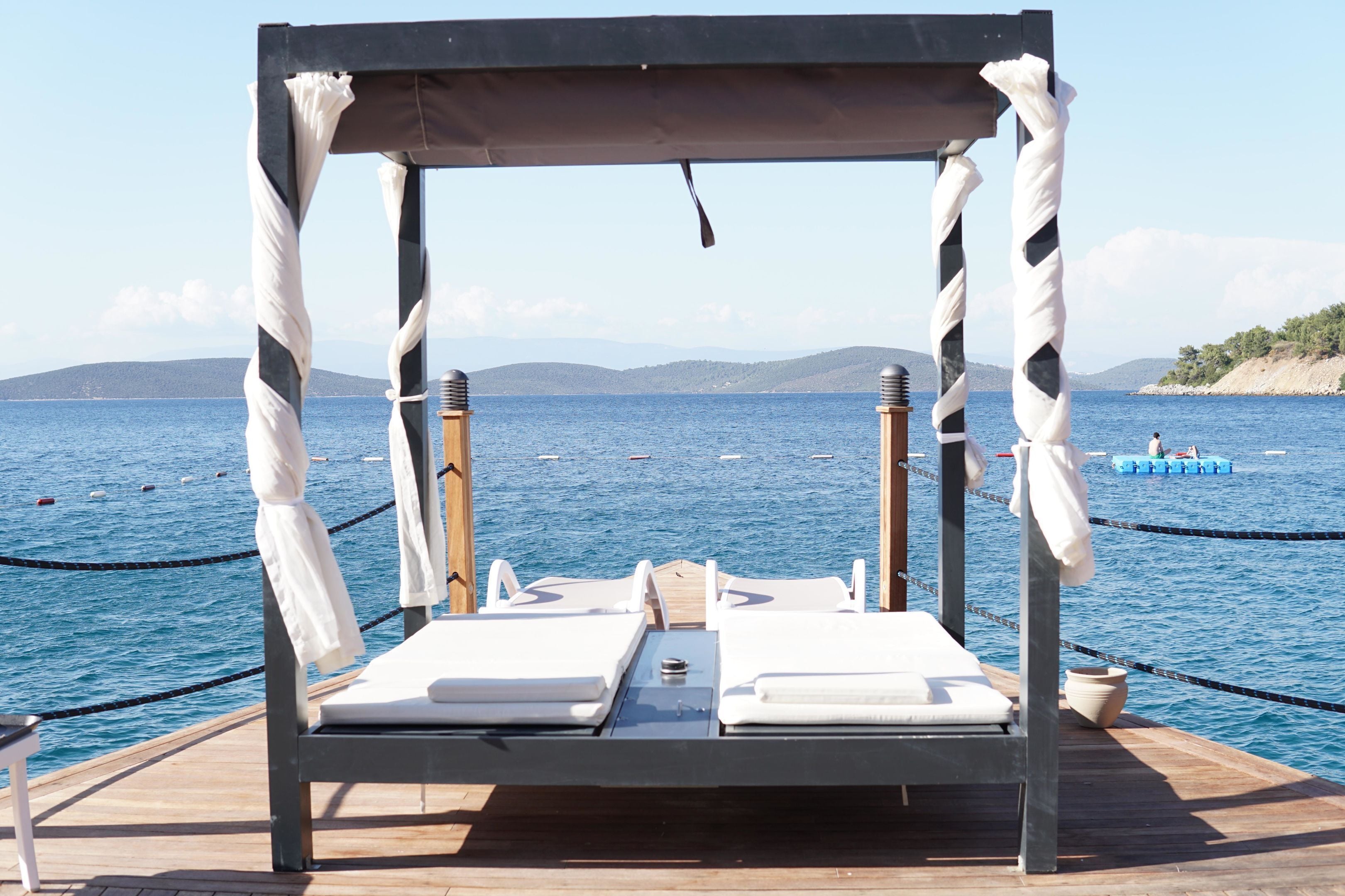 A wooden deck with two white lounge chairs and a canopy, overlooking a calm sea with boats.