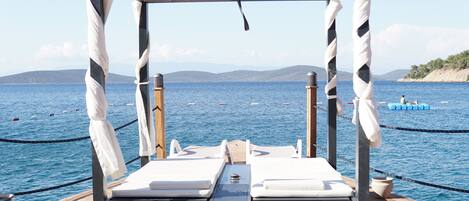 A wooden deck with two white lounge chairs and a canopy, overlooking a calm sea with boats.