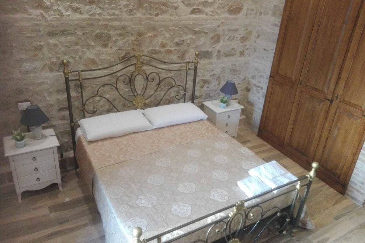 Een slaapkamer met een stenen muur, een bed met metalen frame, een houten garderobe en een nachtkastje met een lamp.