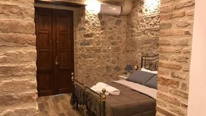 A bedroom with stone walls, a wooden door, a bed with a headboard, and a nightstand with a lamp.