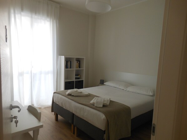 A bedroom with a bed, bedside table, and a small desk with a chair. There is a bookshelf against the wall and a window with curtains.