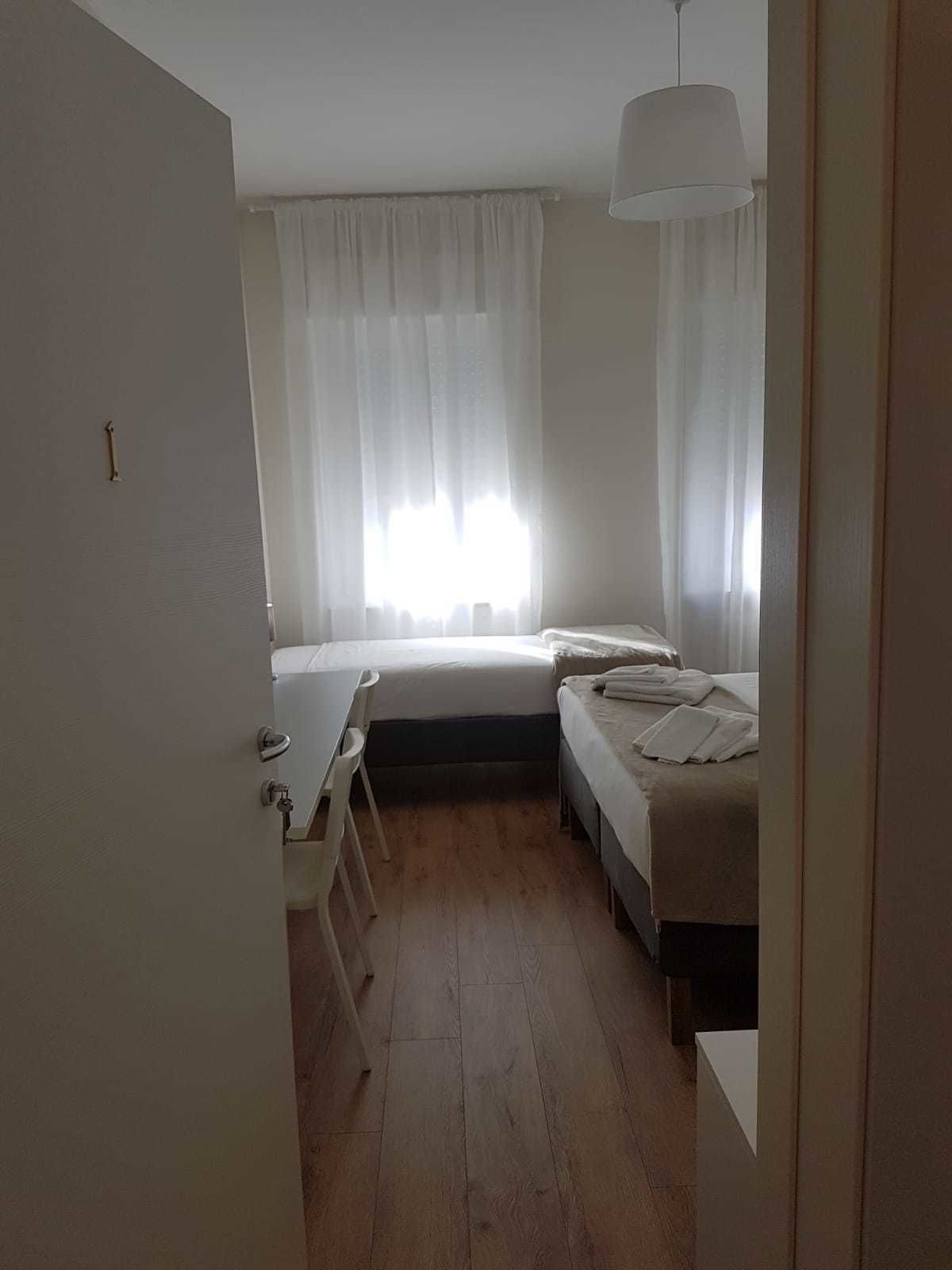 A small, bright room with a bed, a desk, and a chair.