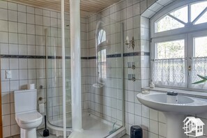 A bathroom with white tiled walls, a glass shower enclosure, a toilet, a sink, and a window with a view of greenery outside. - Cozy, comfortable country house in a dream location (Templin)