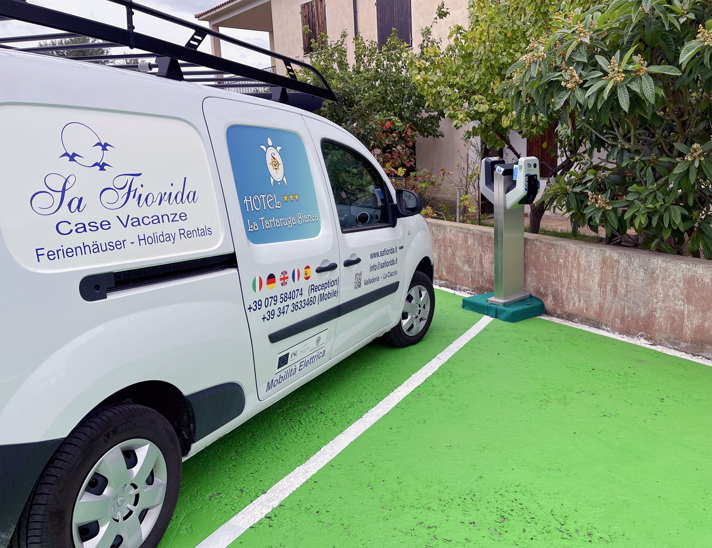 A white van with "La Florida Case Vacanze" and "Holiday Rentals" text, parked on a green surface with a building and trees in the background.