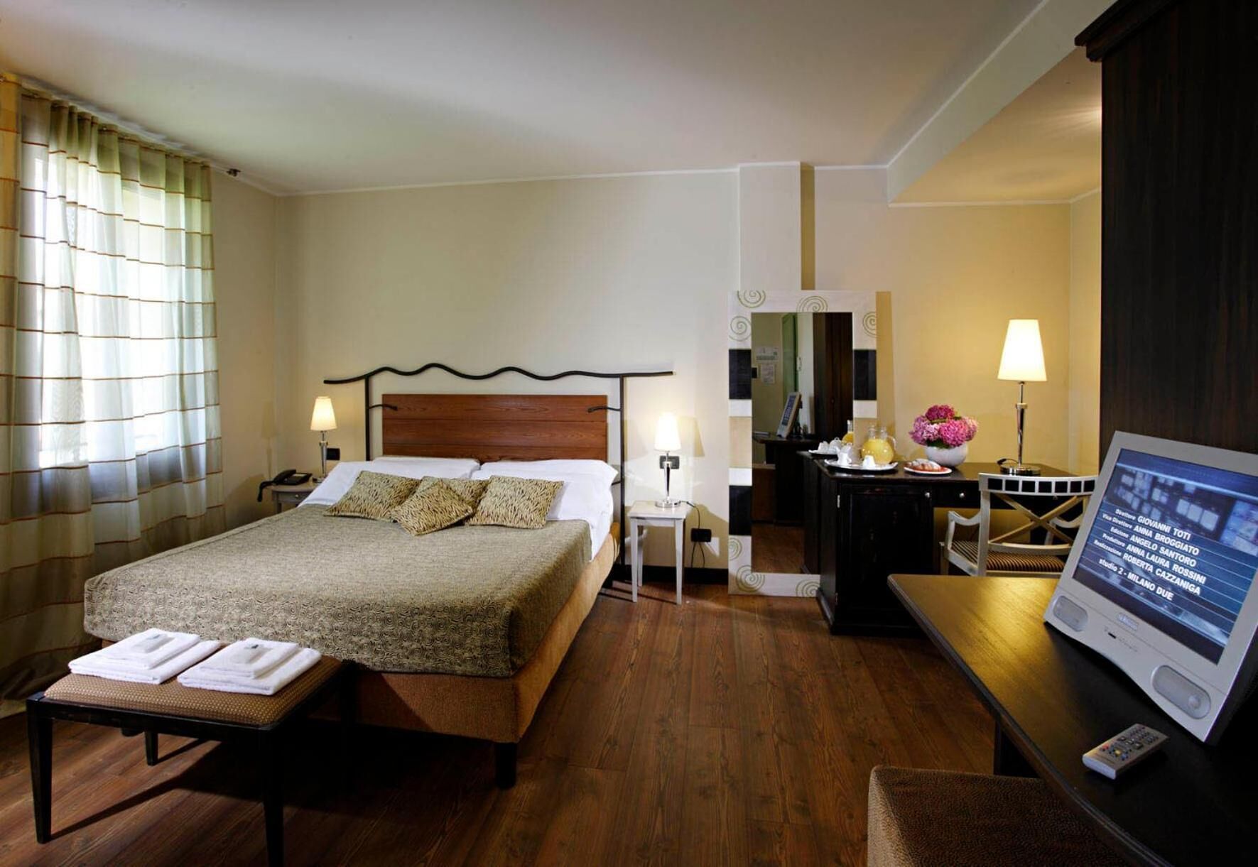 A hotel room with a bed, bedside tables, a desk with a computer, a mirror, and a window with curtains.