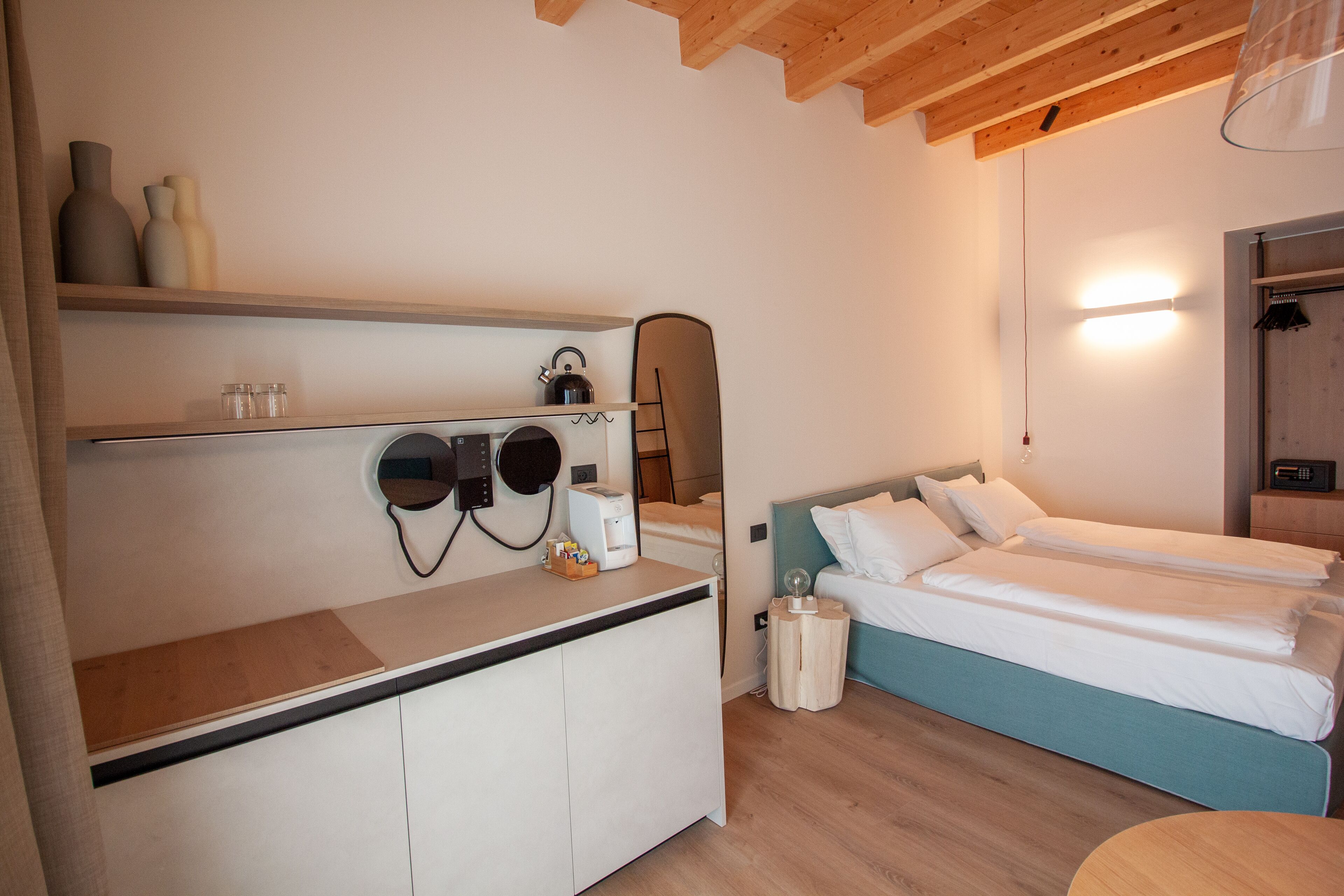 A modern hotel room with a wooden ceiling, a bed with white bedding, a small desk with a kettle, and a mirror above the bed.