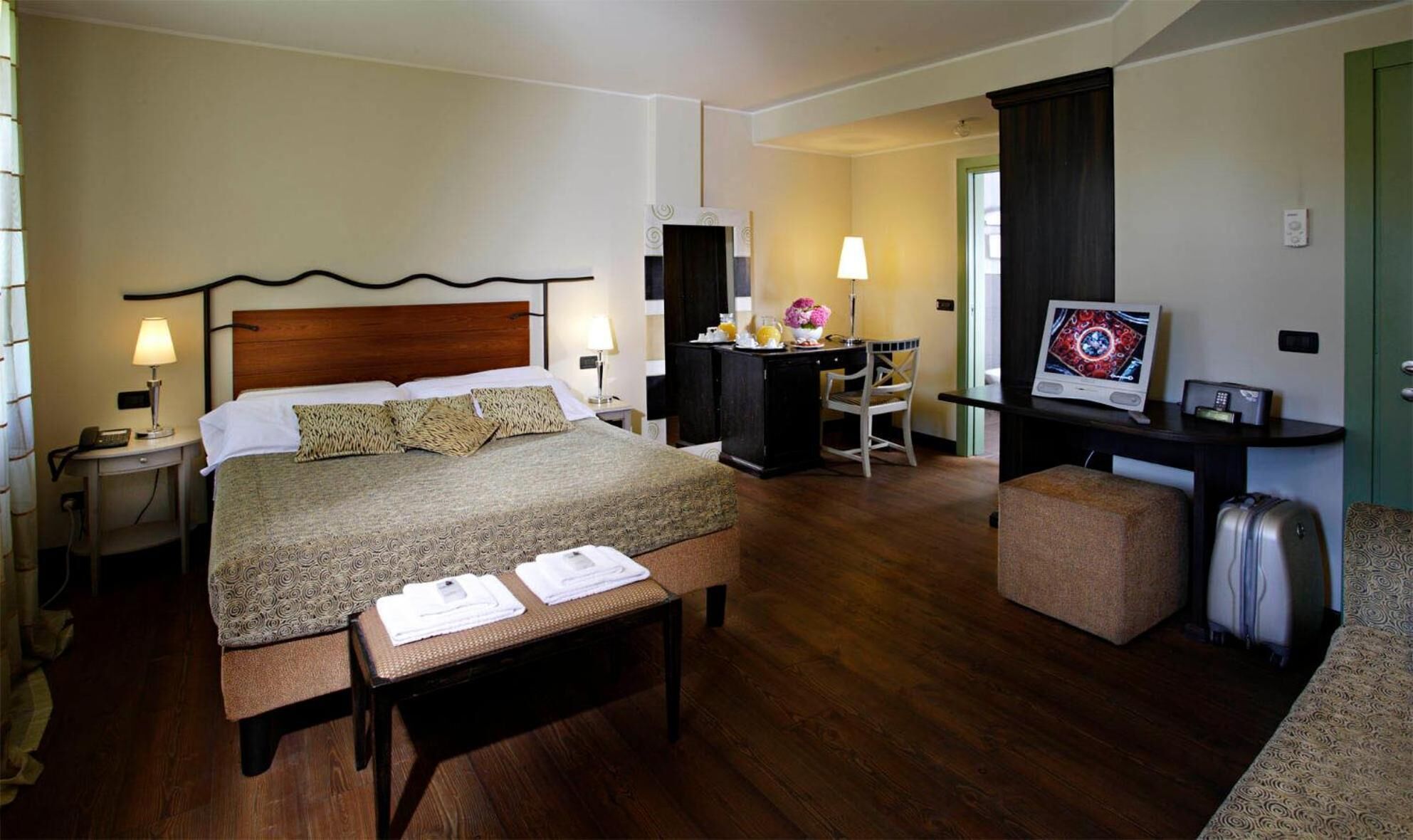 A hotel room with a bed, bedside tables, a desk, a chair, a TV, and a suitcase.