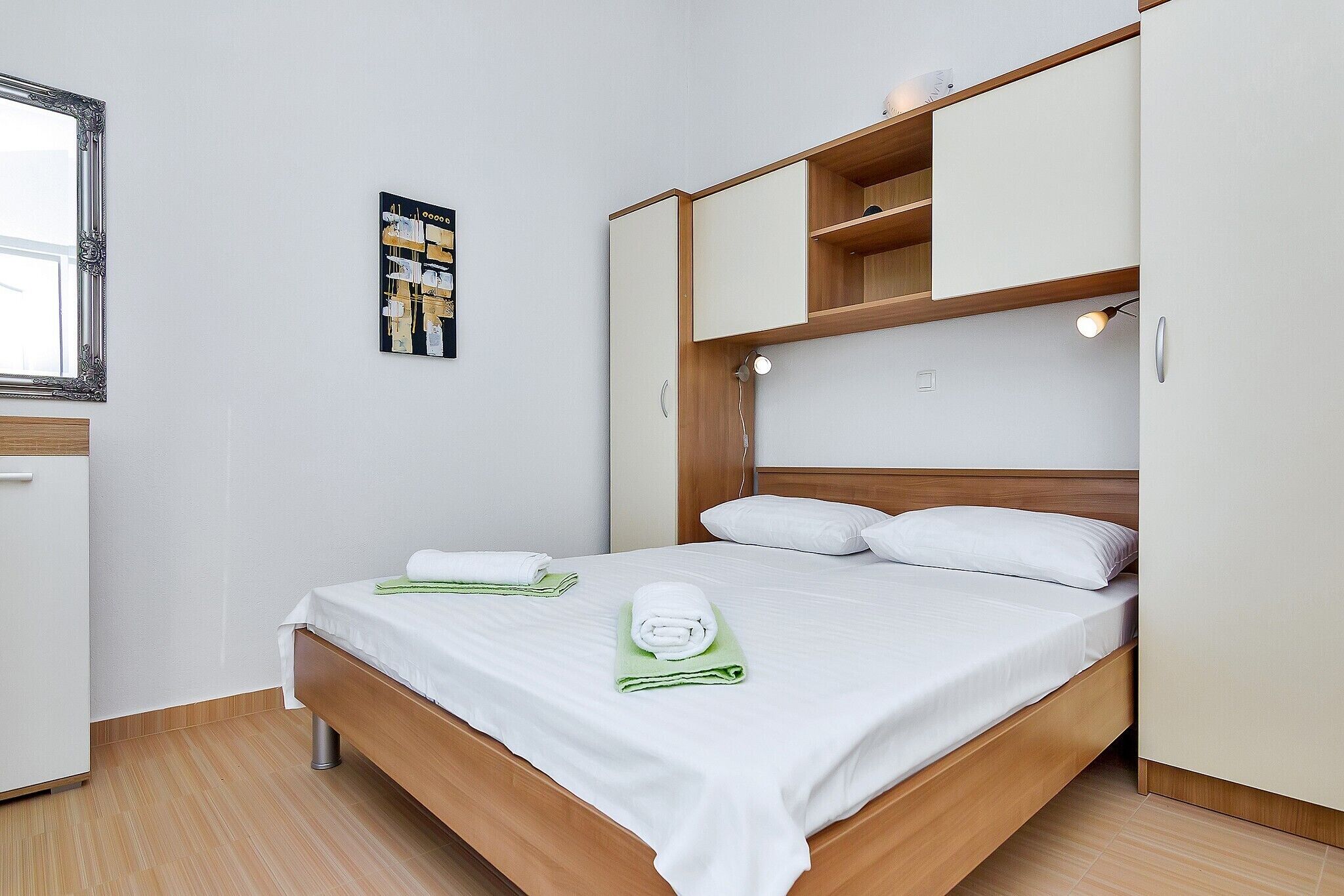 A neatly made bed with white linens and green towels, a wooden headboard, and a small shelf above it.
