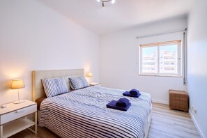 Ein Schlafzimmer mit Bett, Nachttischen, einer Lampe, einem Fenster und einem Korbwäsche.